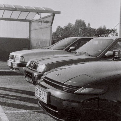 Black and white photograph showing three cars parked in a row outdoors near a covered structure, with the front car in sharp focus and the others receding into the background.