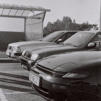 Black and white photograph showing three cars parked in a row outdoors near a covered structure, with the front car in sharp focus and the others receding into the background.