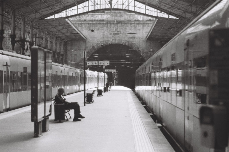 Black and white photograph of a train station platform with two trains on either side, a seated person on a bench near the edge, and a large arched station interior overhead.