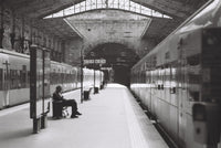 Black and white photograph of a train station platform with two trains on either side, a seated person on a bench near the edge, and a large arched station interior overhead.