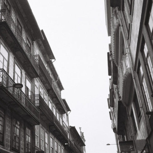 Black and white photograph of narrow European-style buildings with balconies, viewed from below looking up at the sky between the structures.