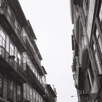 Black and white photograph of narrow European-style buildings with balconies, viewed from below looking up at the sky between the structures.