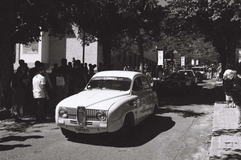 Black and white photograph of a vintage white car parked on a tree-lined street with onlookers standing along the sidewalk.