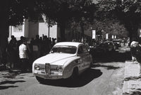 Black and white photograph of a vintage white car parked on a tree-lined street with onlookers standing along the sidewalk.
