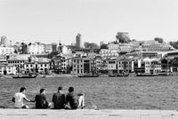 Black and white photograph of a waterfront cityscape with several people sitting on a stone embankment in the foreground, looking toward a row of buildings across the water.