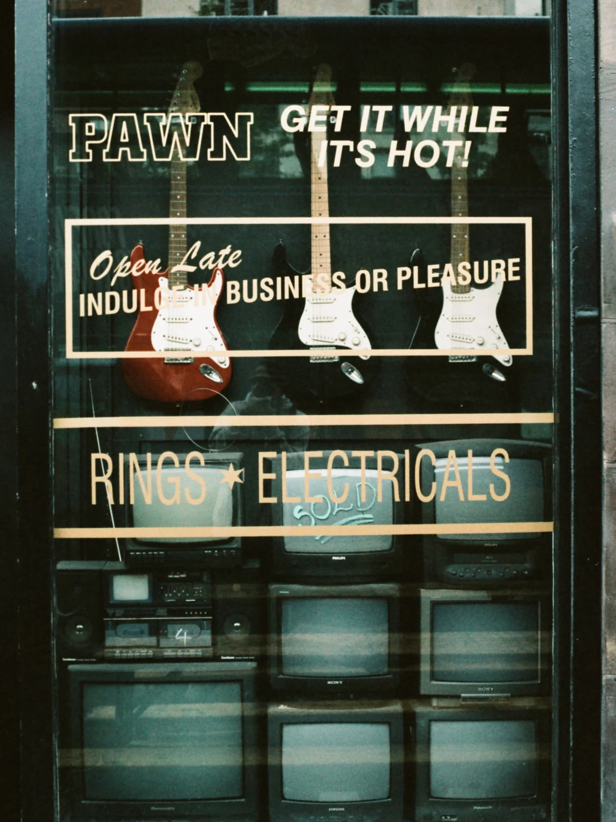 Vintage storefront window display with electric guitars hanging behind glass and multiple CRT television sets below. Large window decals read 'PAWN GET IT WHILE IT'S HOT!' and 'RINGS * ELECTRICALS'.