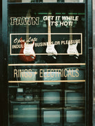 Vintage storefront window display with electric guitars hanging behind glass and multiple CRT television sets below. Large window decals read 'PAWN GET IT WHILE IT'S HOT!' and 'RINGS * ELECTRICALS'.