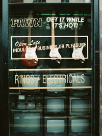 Vintage storefront window display with electric guitars hanging behind glass and multiple CRT television sets below. Large window decals read 'PAWN GET IT WHILE IT'S HOT!' and 'RINGS * ELECTRICALS'.