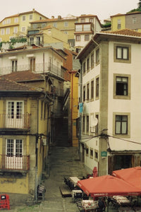 A narrow cobblestone alleyway between old European-style buildings with cream, beige, and ochre facades, terracotta roofs, outdoor cafe seating with red umbrellas, and a staircase rising up the center.