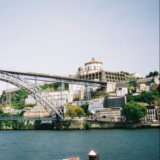 A scenic photograph of a large steel arch bridge spanning a river, with buildings and green hillsides on the right bank under a pale sky.