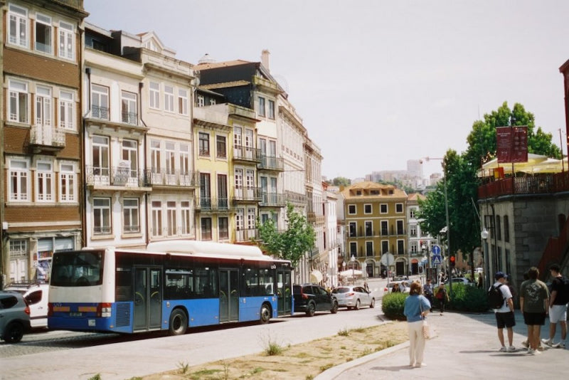 A city street scene with a blue and white bus driving past multi-story residential buildings, with pedestrians on a sidewalk under a bright sky.