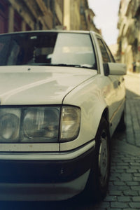 A vintage white Mercedes-Benz sedan parked on a cobblestone street with blurred buildings in the background.