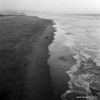 Two people walking along a beach with waves and a distant landscape.
