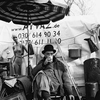 Man sitting under an umbrella with a cup, surrounded by various items and a banner with contact information.