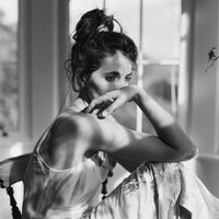 A black and white fashion photograph of a woman in a light satin slip dress, sitting on a chair in a brightly lit room with large windows.