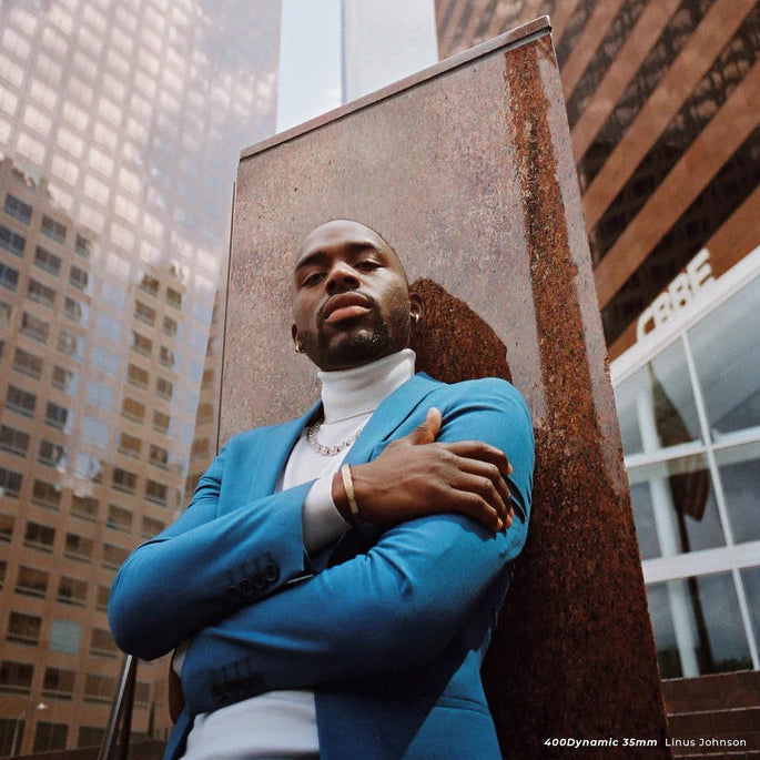 A man wearing a bright blue blazer over a white turtleneck stands with arms crossed in front of a rusted metal sculpture, with tall city buildings reflected in the glass behind him.