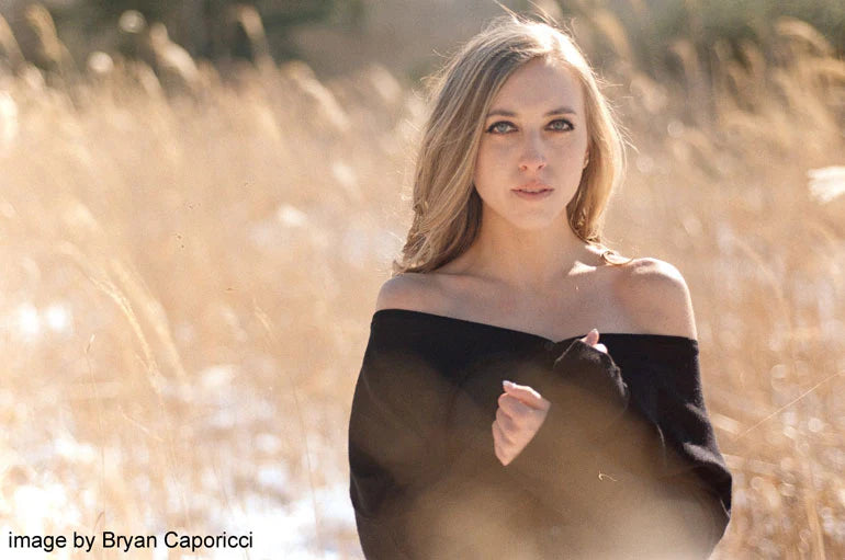 A woman with blonde hair wearing a black off-the-shoulder top stands in a field of dry golden grass, photographed in warm natural light with a soft blurred background.