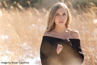 A woman with blonde hair wearing a black off-the-shoulder top stands in a field of dry golden grass, photographed in warm natural light with a soft blurred background.