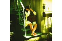 A woman in a white dress sits barefoot on outdoor stairs holding a dark acoustic guitar, with a green-tinted warm light illuminating the scene.