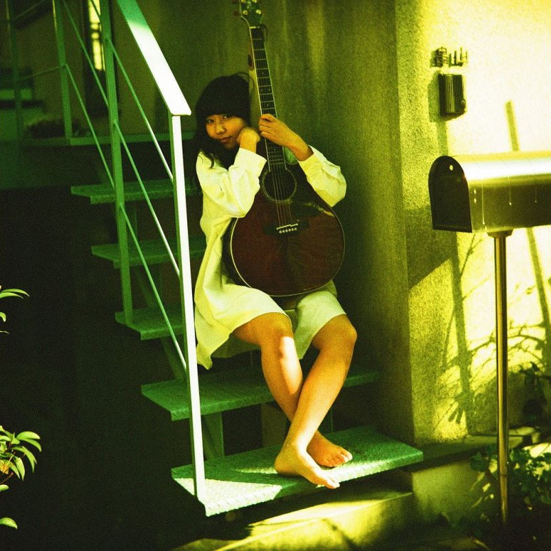 A woman in a white dress sits barefoot on outdoor stairs holding a dark acoustic guitar, with a green-tinted warm light illuminating the scene.