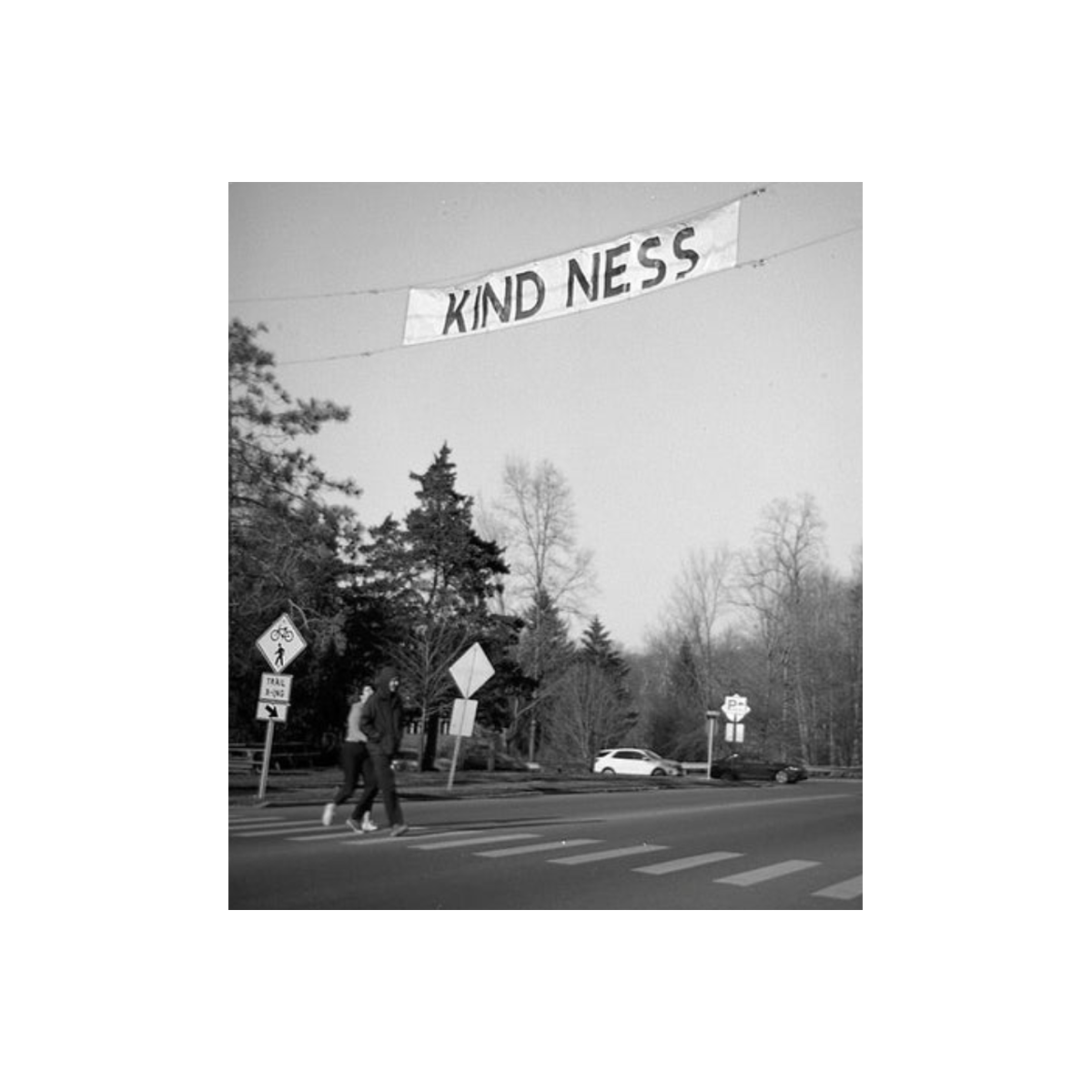 Black and white 620 film shot of a street scene with a banner reading KIND NESS hanging above a crosswalk.