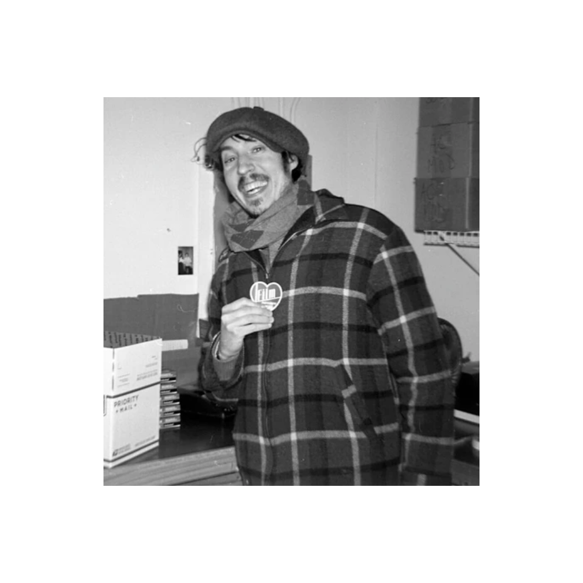 Smiling man wearing a plaid jacket and cap, holding a heart-shaped Film Photography Project pin in a studio setting.