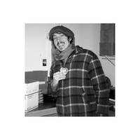 Smiling man wearing a plaid jacket and cap, holding a heart-shaped Film Photography Project pin in a studio setting.