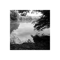Black and white landscape photograph of a calm lake or river bordered by dense trees, with reflections in the water and dark foliage framing the scene.