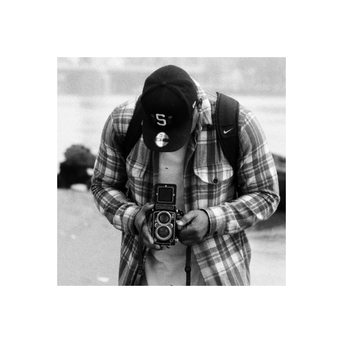 A black and white lifestyle photograph of a man wearing a plaid flannel shirt, a baseball cap with an 'S' logo, and a camera backpack, holding a vintage twin-lens reflex camera.
