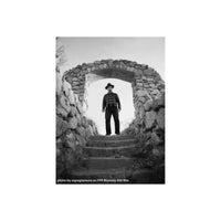 Black and white photograph of a man wearing a hat, striped shirt, and dark trousers standing on stone steps within a rustic stone archway.