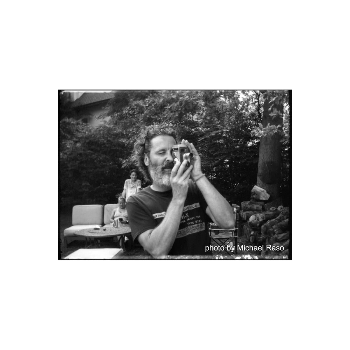 Man with a beard holding a vintage camera in a high-contrast, black and white outdoor scene shot on FPP Sonic 25 film.