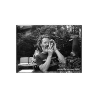Man with a beard holding a vintage camera in a high-contrast, black and white outdoor scene shot on FPP Sonic 25 film.
