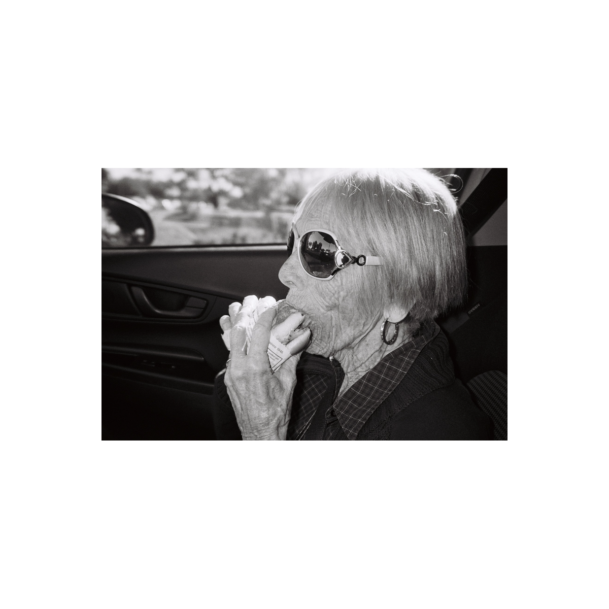 Black and white film shot of an elderly woman wearing sunglasses eating a sandwich inside a car.