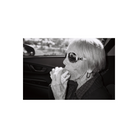 Black and white film shot of an elderly woman wearing sunglasses eating a sandwich inside a car.