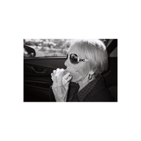Black and white film shot of an elderly woman wearing sunglasses eating a sandwich inside a car.