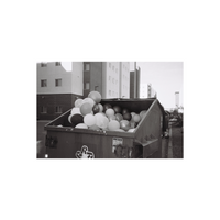 Black and white film photograph of a metal dumpster filled with a large pile of round balloons outside an apartment building.