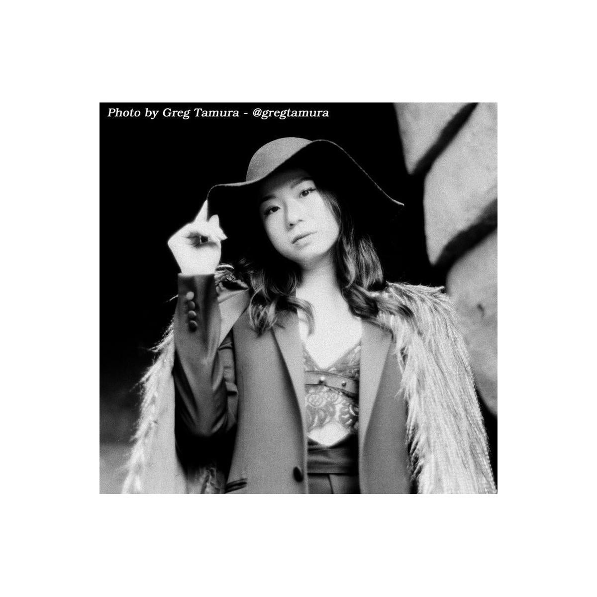 Black and white portrait of a woman wearing a wide-brimmed hat, a patterned top, and a textured jacket with a shawl collar, posed against a dark background. Text at the top reads 'Photo by Greg Tamura - @gregtamura'.