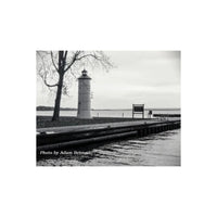 Black and white photograph of a lighthouse beside a body of water, with a tree on the left, a wooden dock in the foreground, and a cloudy sky overhead. Text at the bottom reads 'Photo by Adam Bennett'.