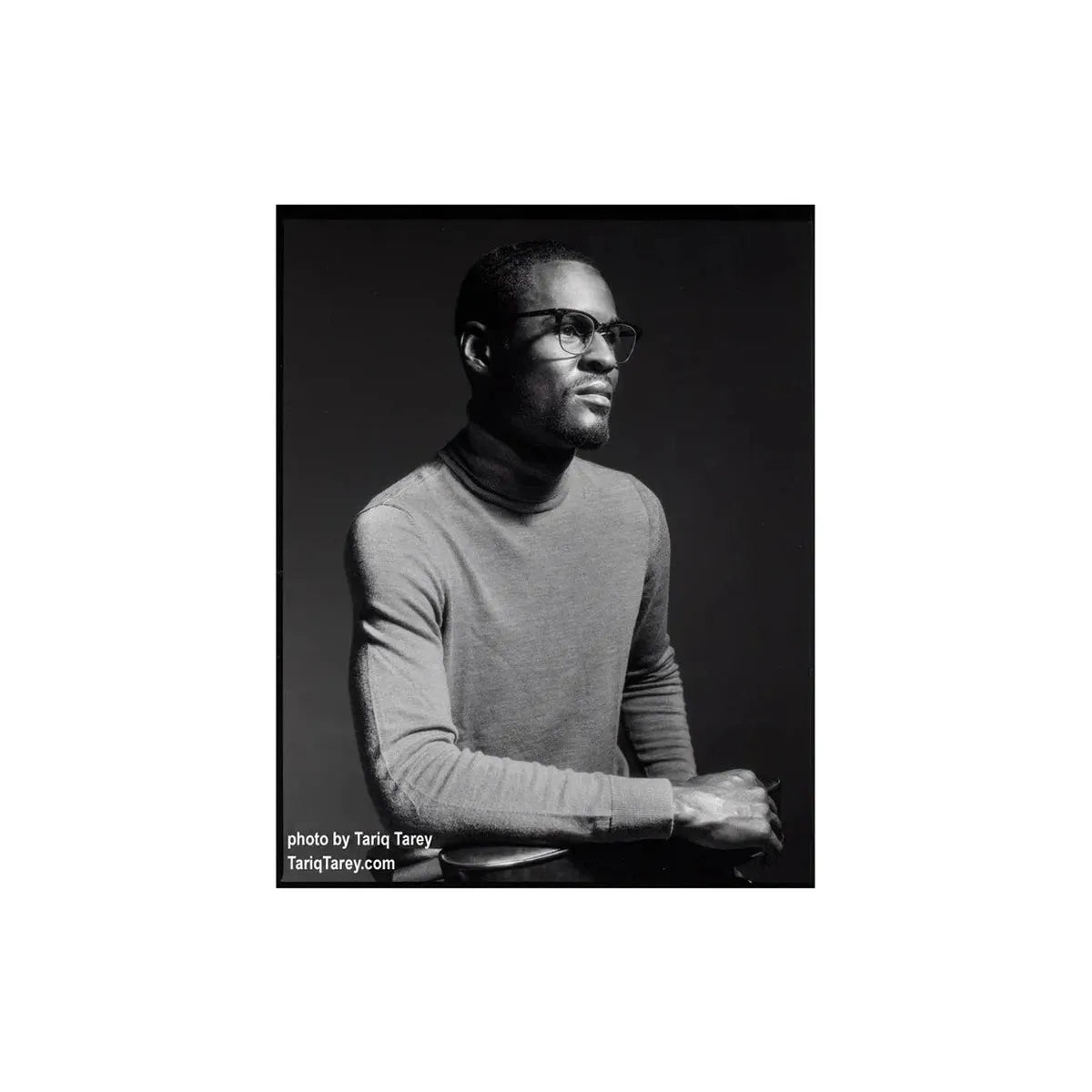 A black and white portrait photograph of a man wearing thick-rimmed glasses and a gray turtleneck sweater, seated against a dark studio background. White text in the lower-left corner reads 'photo by Tariq Tarey'.