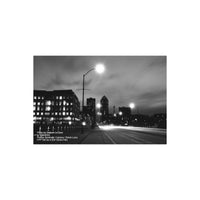 Black and white long-exposure city street scene at night with illuminated buildings, a lit streetlamp, road light trails, and cloudy sky. Small photo credits are printed in the lower left corner.