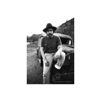 A black and white photograph of a man wearing a wide-brimmed hat, a dark t-shirt, and light-colored jeans, sitting on the front of a vintage truck in an outdoor, rugged landscape.