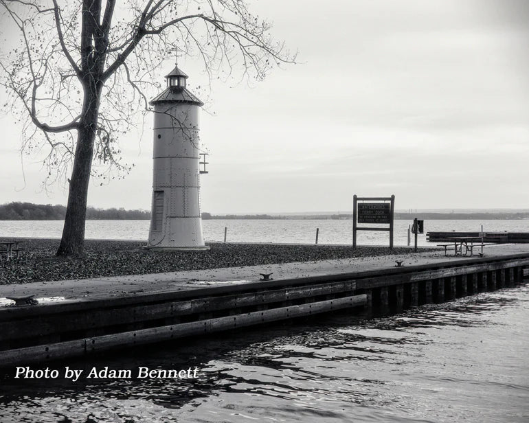 Black and white photograph of a lighthouse beside a body of water, with a tree on the left, a wooden dock in the foreground, and a cloudy sky overhead. Text at the bottom reads 'Photo by Adam Bennett'.