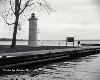 Black and white photograph of a lighthouse beside a body of water, with a tree on the left, a wooden dock in the foreground, and a cloudy sky overhead. Text at the bottom reads 'Photo by Adam Bennett'.