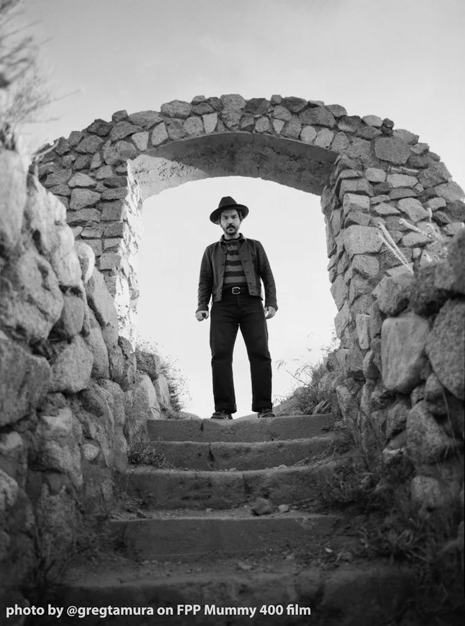 Black and white photograph of a man wearing a hat, striped shirt, and dark trousers standing on stone steps within a rustic stone archway.