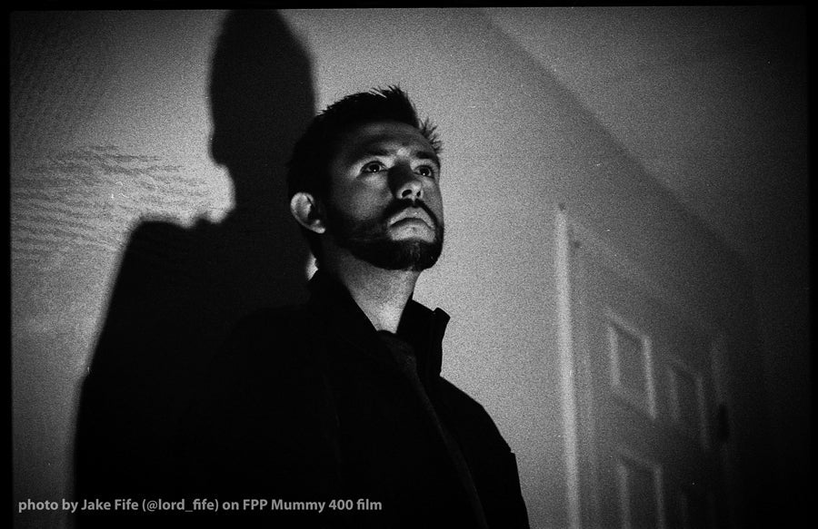 Black and white portrait photograph of a man looking upward, with a dramatic shadow cast on the wall behind him. Small text at the bottom reads 'photo by Jake Fife (@lord_fife) on FPP Mummy 400 film'.