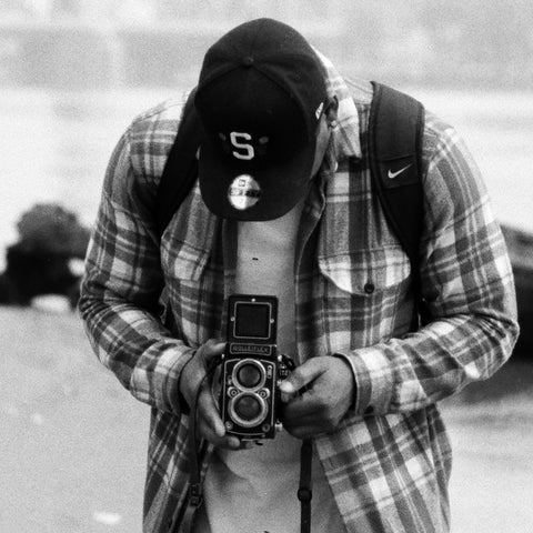 A black and white lifestyle photograph of a man wearing a plaid flannel shirt, a baseball cap with an 'S' logo, and a camera backpack, holding a vintage twin-lens reflex camera.