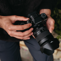 A close-up of a person holding a black Fujifilm mirrorless camera with a lens attached, shown in a real-world setting with hands visible and a blurred background.