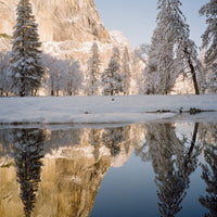A winter landscape photograph of a snow-covered granite cliff and trees reflected in a calm dark pool of water under a pale blue sky.