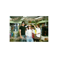 Three people standing in a convenience store aisle, with a decorative translucent tribal pattern overlaid across the entire image. The person on the left wears a black t-shirt and jeans.