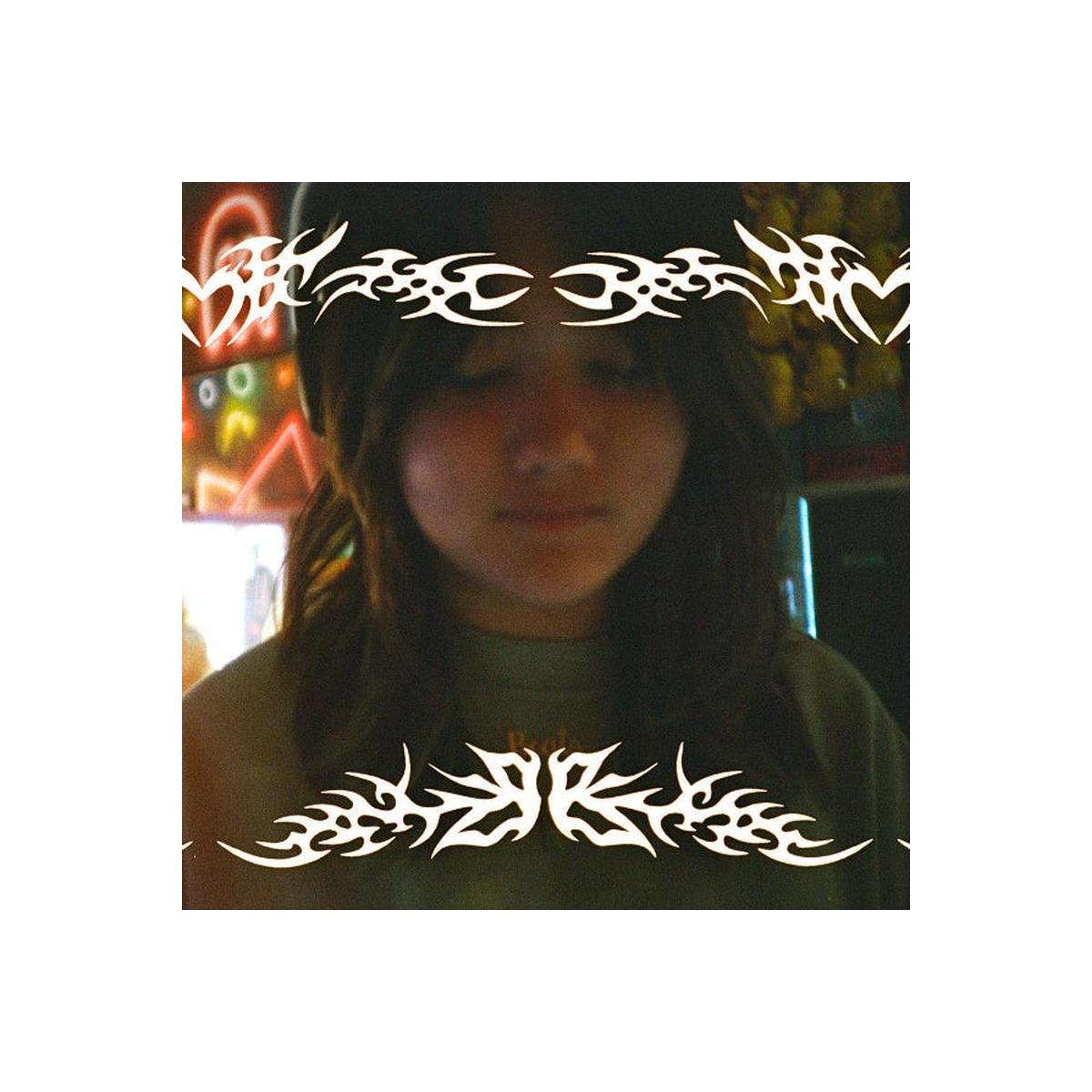 A person with long dark hair wearing a dark green t-shirt, standing in a brightly lit arcade or game center with colorful neon lights in the background. The image has a heavy white tribal graphic overlay on the top and bottom borders.
